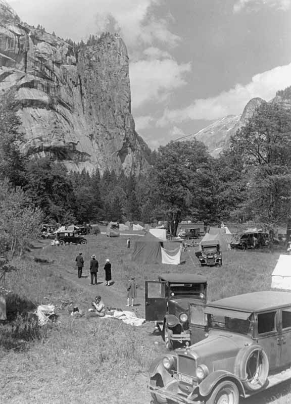 Touring cars and canvas tents scattered across a meadow beneath a sheer granite cliff.