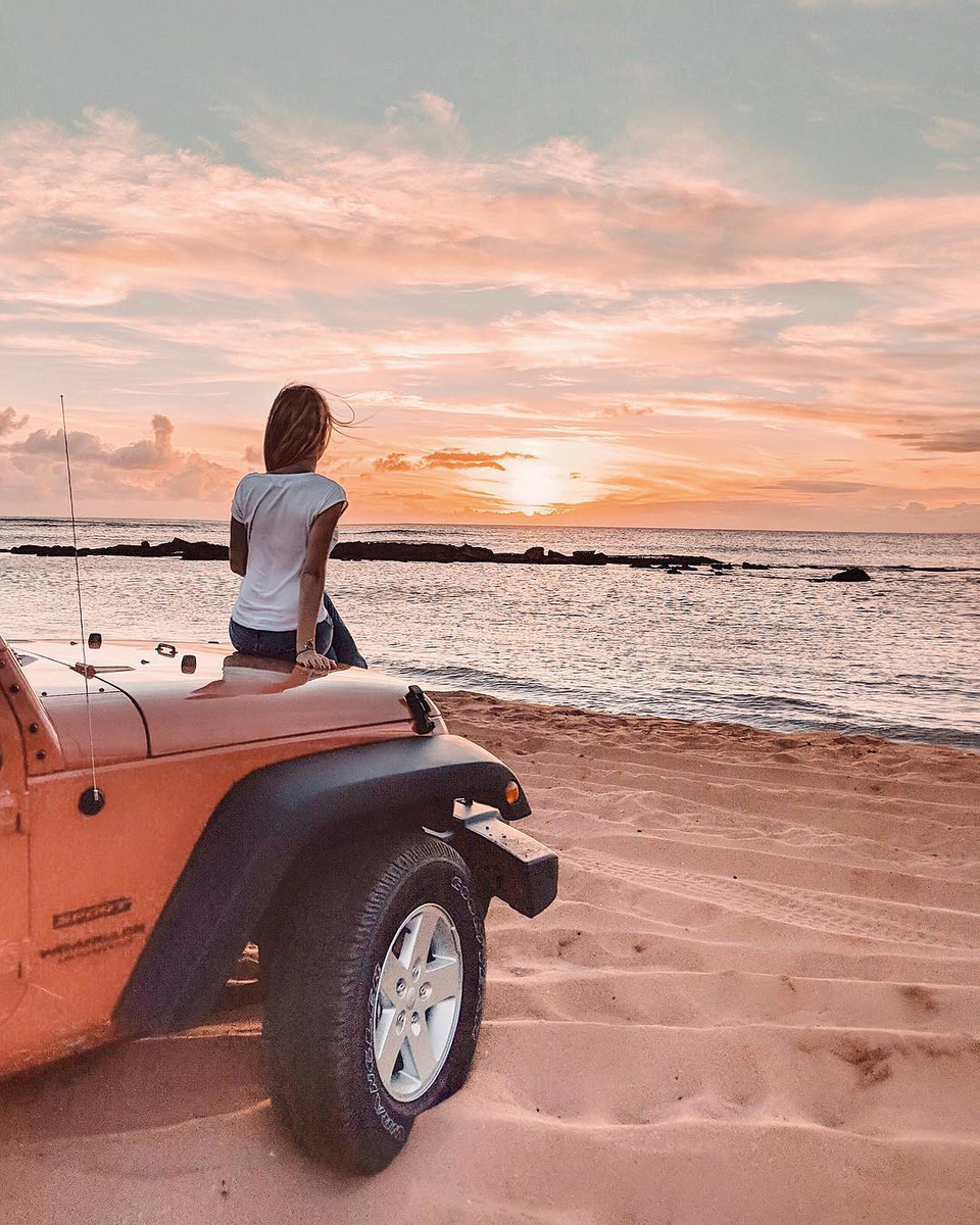 Olga sitting on the bonnet of a red Jeep on a beach, watching the sun set over the sea.