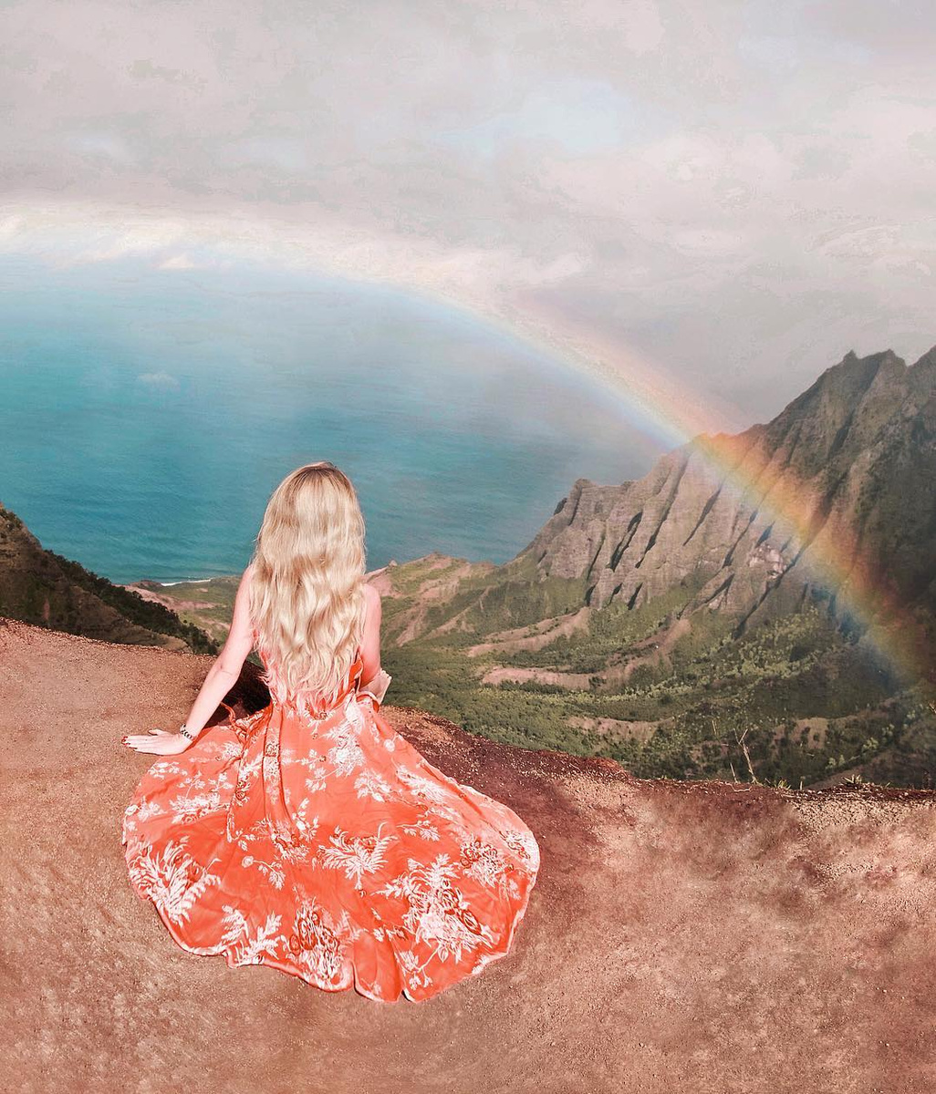 Olga in a long red dress sitting above the Kalalau Valley on Kauaʻi, with a rainbow over the green ridges and the sea.