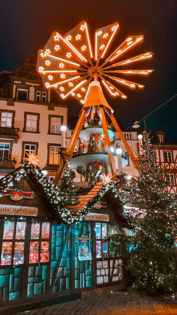 A glowing Christmas pyramid above wooden market stalls and decorated trees at the Trier Christmas market.
