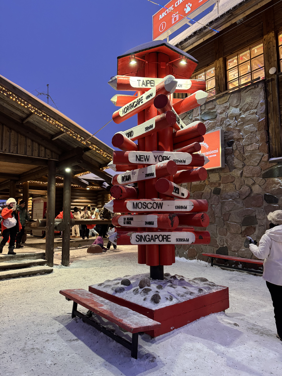 Red wooden signpost pointing towards world cities beside the Arctic Circle information building in Santa Claus Village.