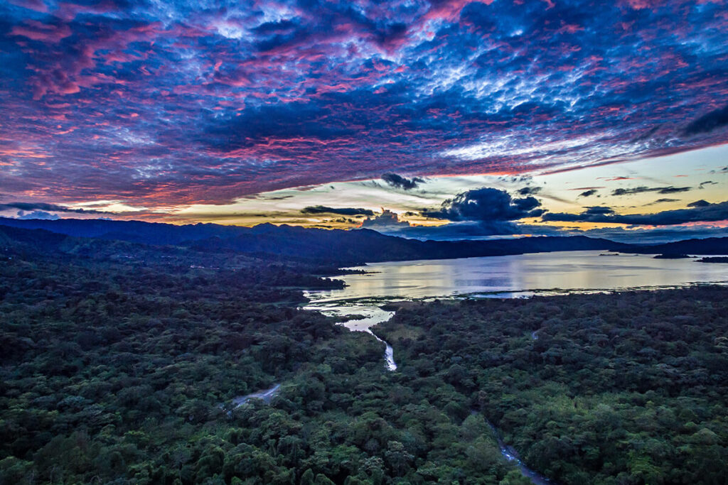 An aerial view across rainforest and a river towards Lake Arenal beneath a coloured evening sky.