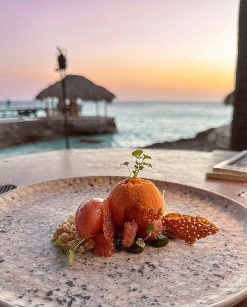 A plated dessert at Brass Boer with the sea and sunset behind it.