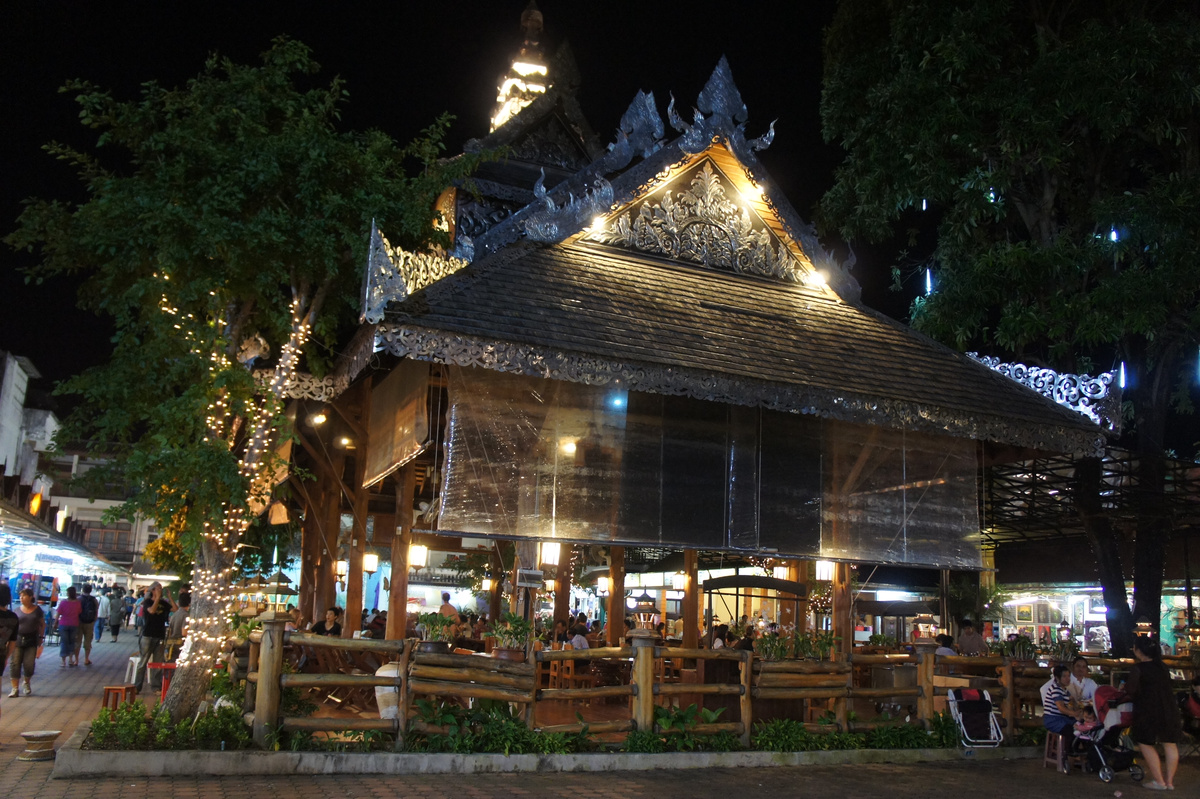 An ornate wooden pavilion glows among trees, food tables and stalls at Chiang Rai’s night market.