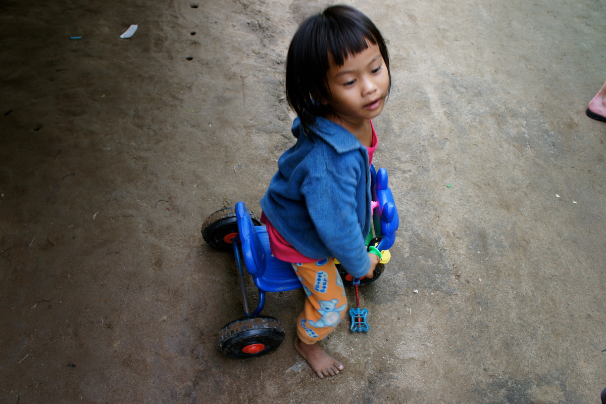 A child in a blue top sitting on a small blue tricycle on a village dirt path.