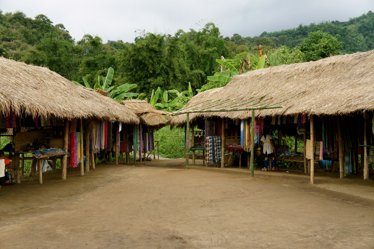 Thatched stalls arranged around an open dirt courtyard with forested hills beyond.