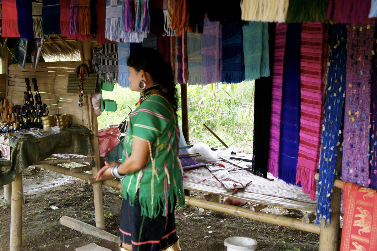 A woman standing beside a loom and hanging textiles inside a thatched stall.