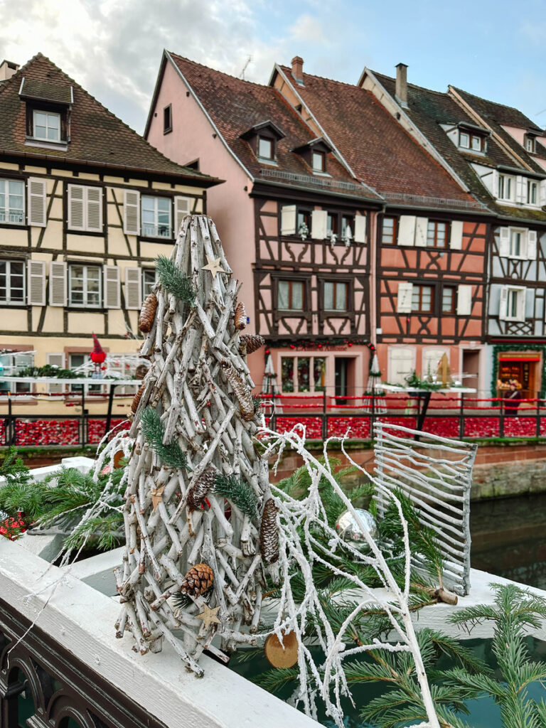 Fir branches and wooden Christmas decorations beside a Colmar canal, with half-timbered houses behind.