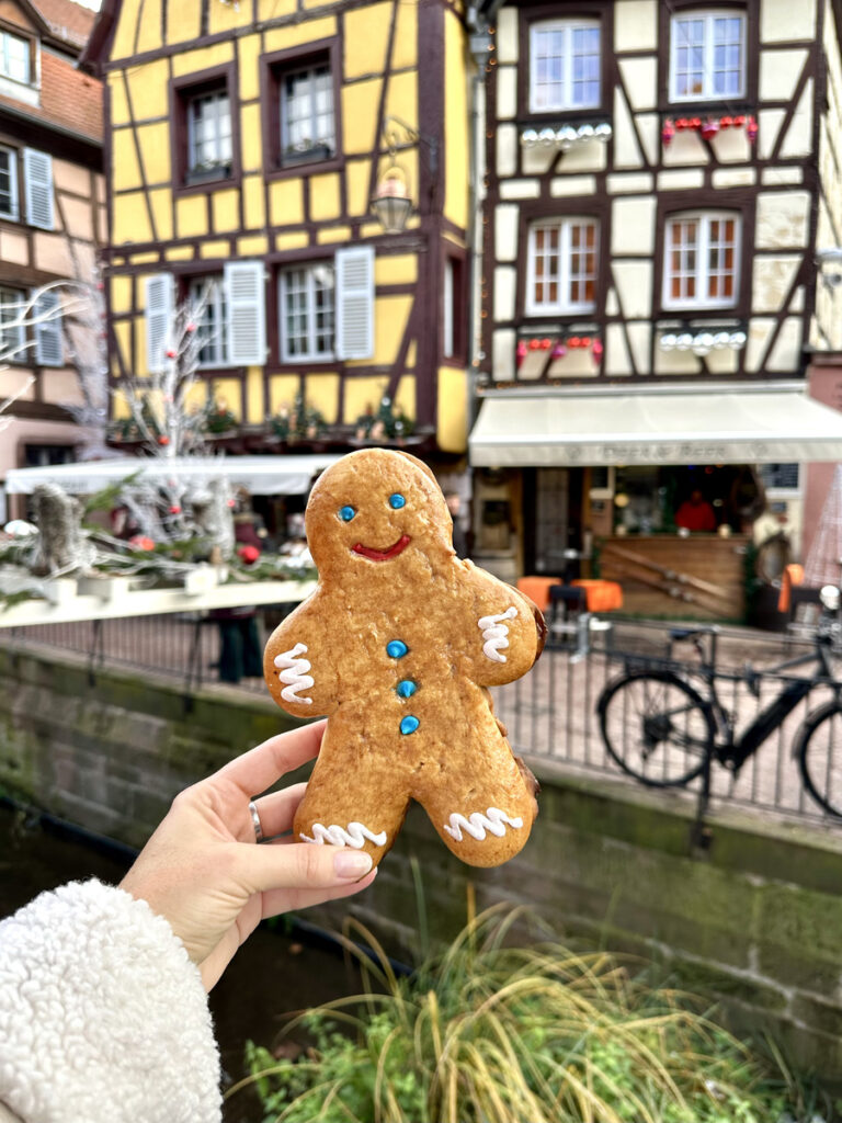 A decorated gingerbread figure held beside a canal and half-timbered houses in Colmar.