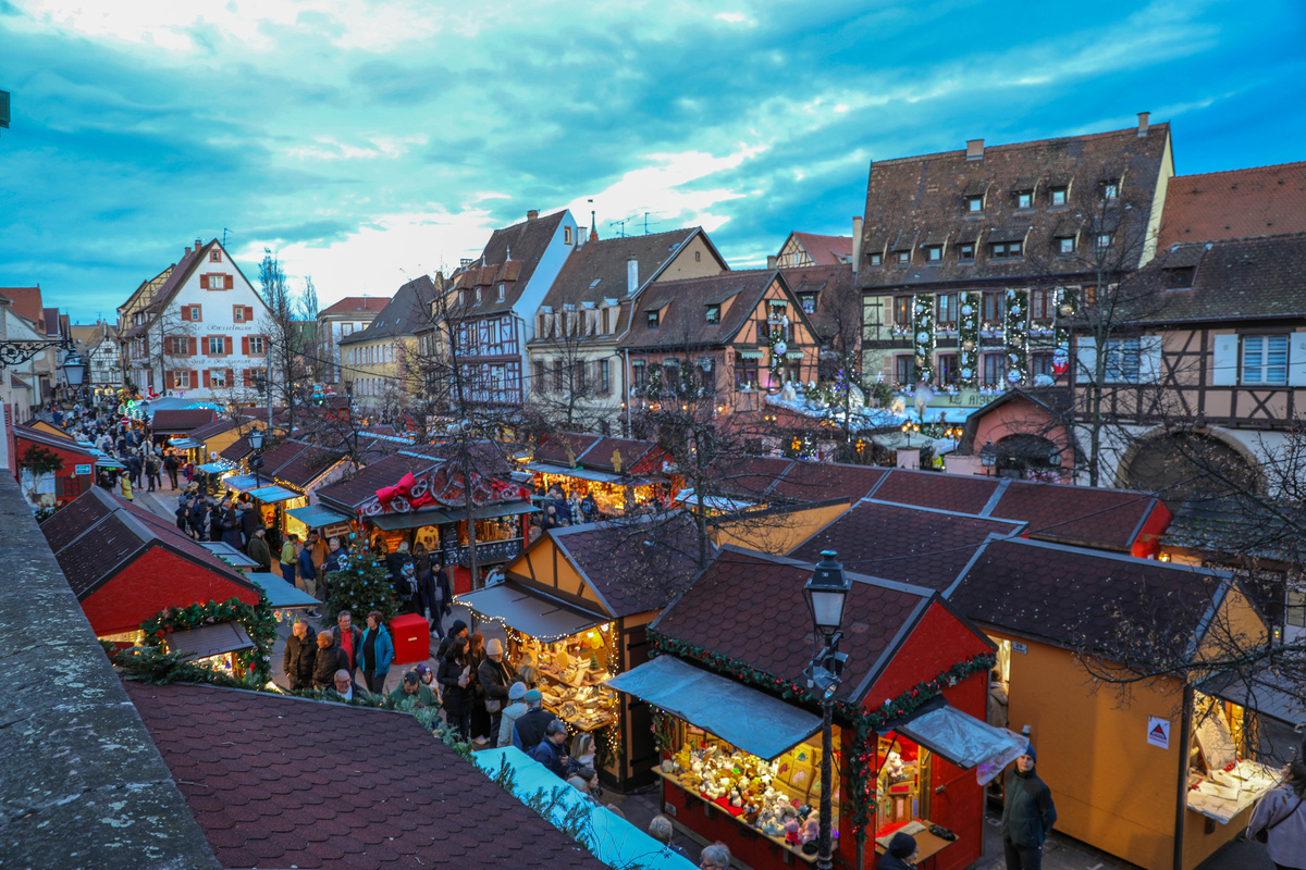 Visitors walk through narrow lanes between Christmas chalets at Colmar’s Place des Six Montagnes Noires, seen from a raised viewpoint.