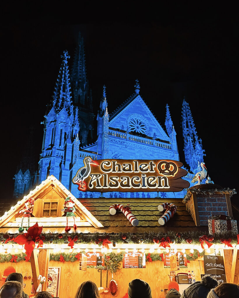 Colmar’s Saint Martin’s Church illuminated blue above the Chalet Alsacien Christmas food stall.