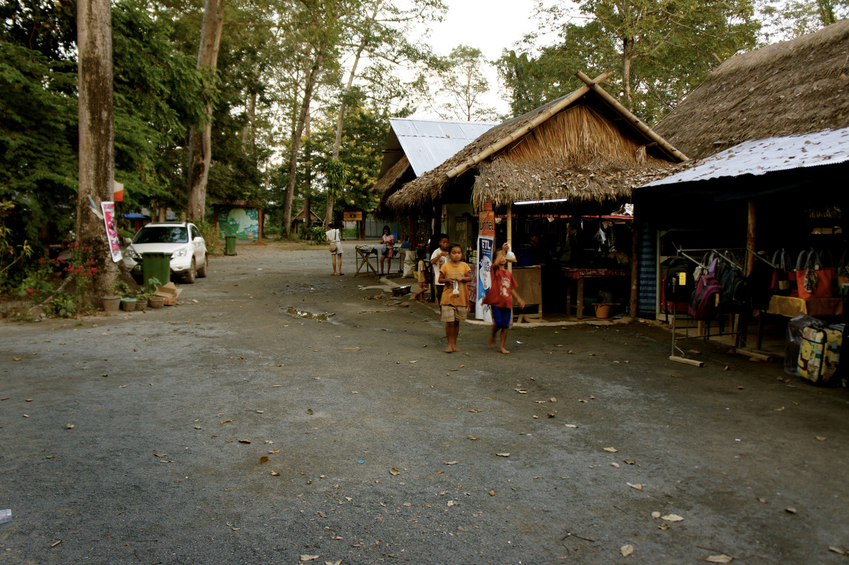 A lane lined with thatched market stalls and bags on display at Don Sao, with children walking past.