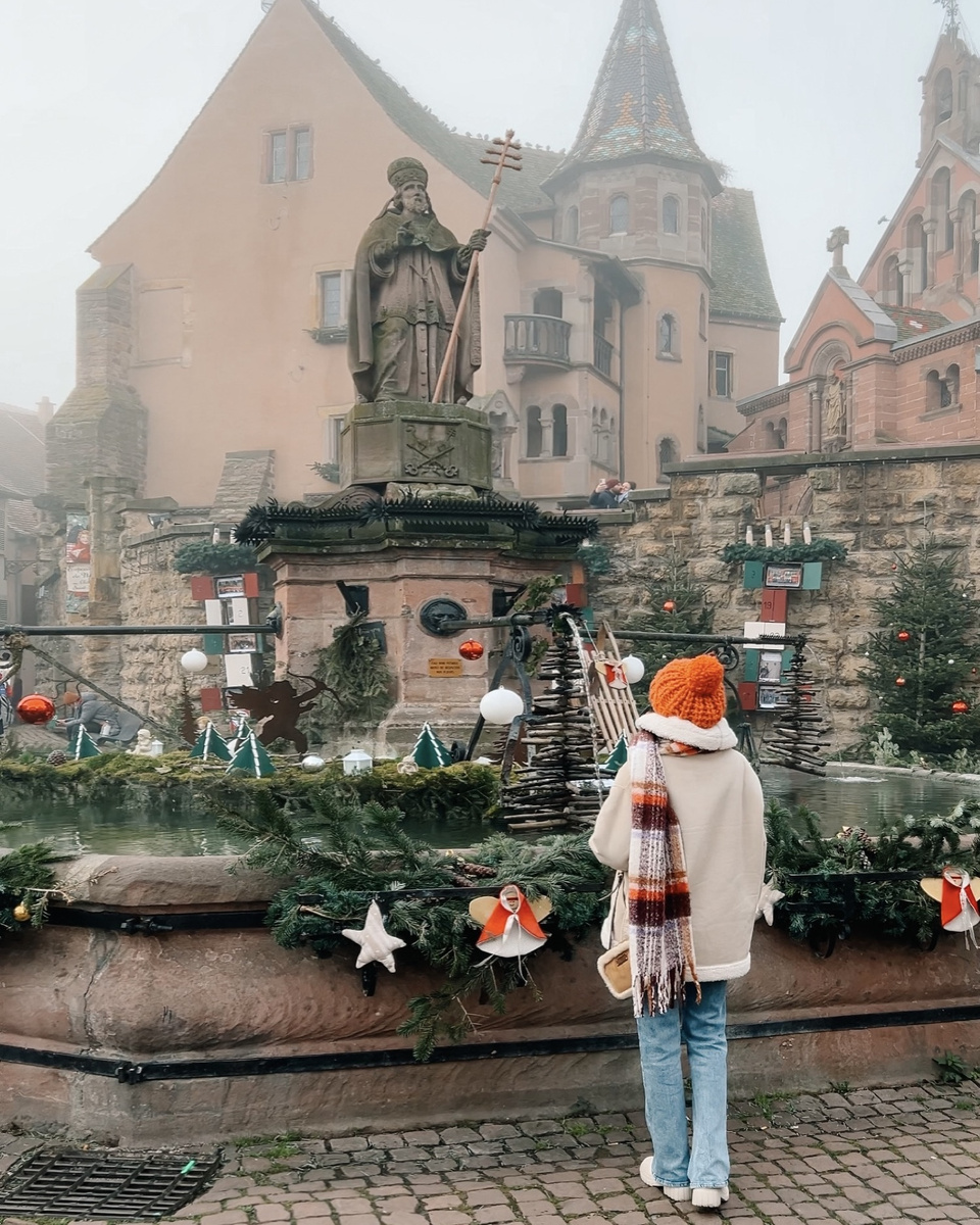 Olga looks towards Eguisheim’s Saint-Léon fountain, surrounded by fir garlands and Christmas decorations.