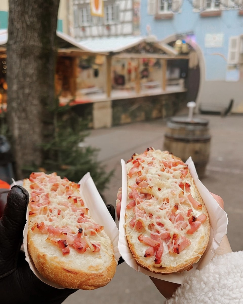 Two toasted bread halves topped with melted cheese and ham held beside Christmas stalls in Eguisheim.