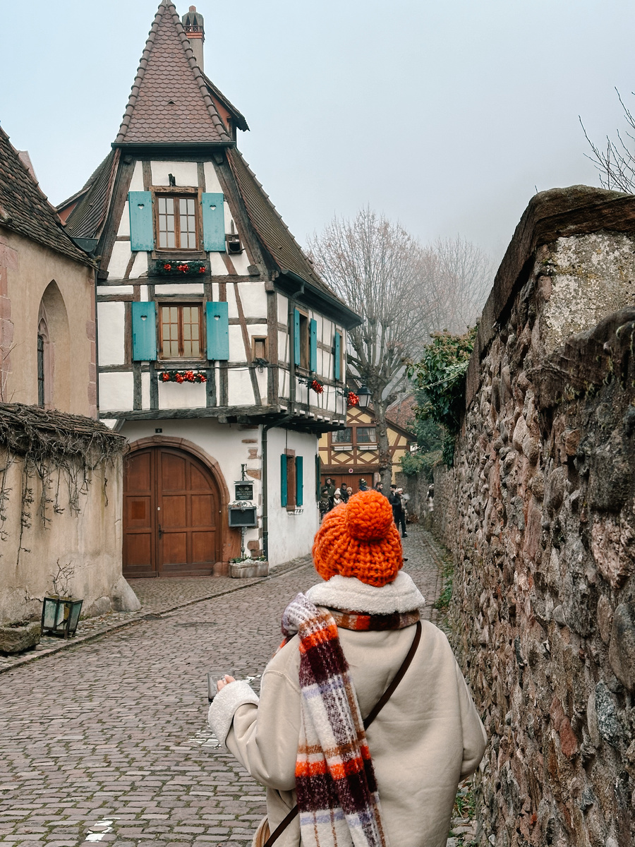 Olga walks along a cobbled Eguisheim lane towards the narrow half-timbered Pigeonnier house with green shutters.