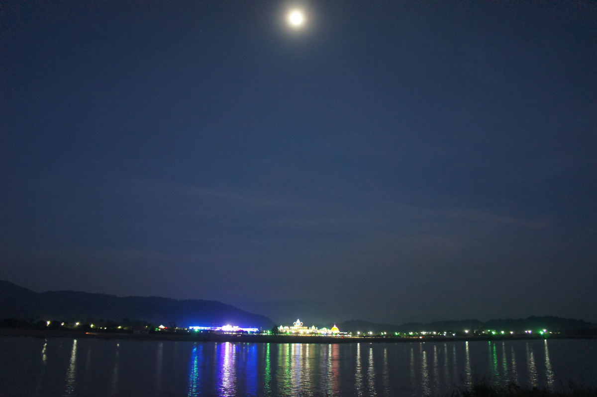 A bright moon hangs above the dark Mekong, with illuminated buildings reflected across the water.