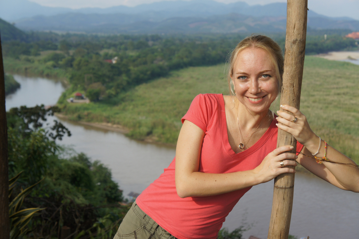 A woman in a red shirt leans against a wooden post above the Ruak and Mekong river junction.
