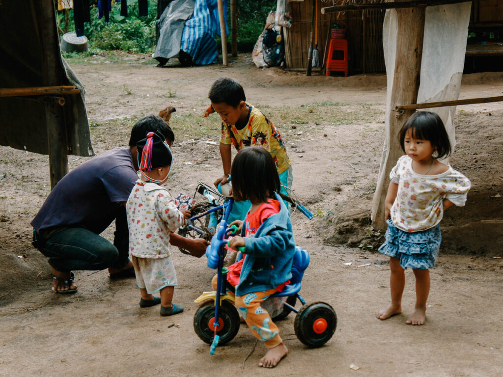 Children gather around a blue tricycle while an adult crouches beside it in a village courtyard.
