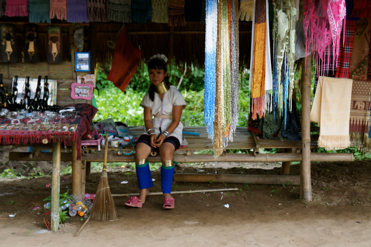 A woman wearing brass neck rings sits among hanging textiles and jewellery at a village stall.
