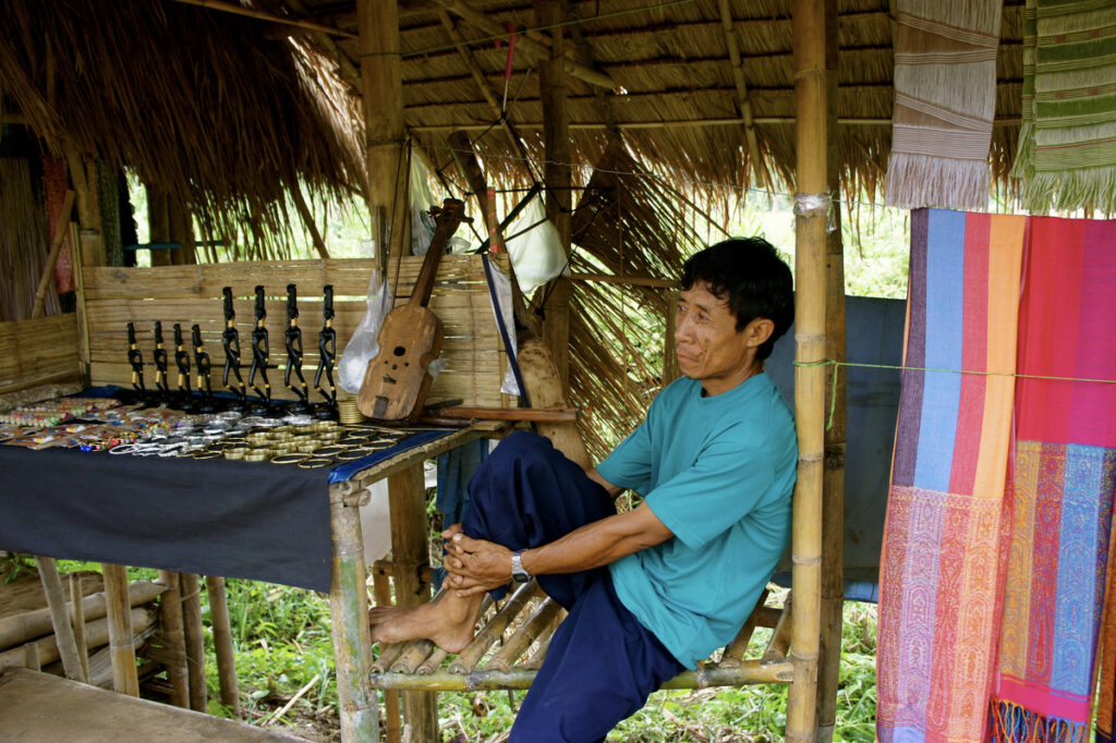 A person sits beside textiles, jewellery and a wooden stringed instrument beneath a thatched roof.