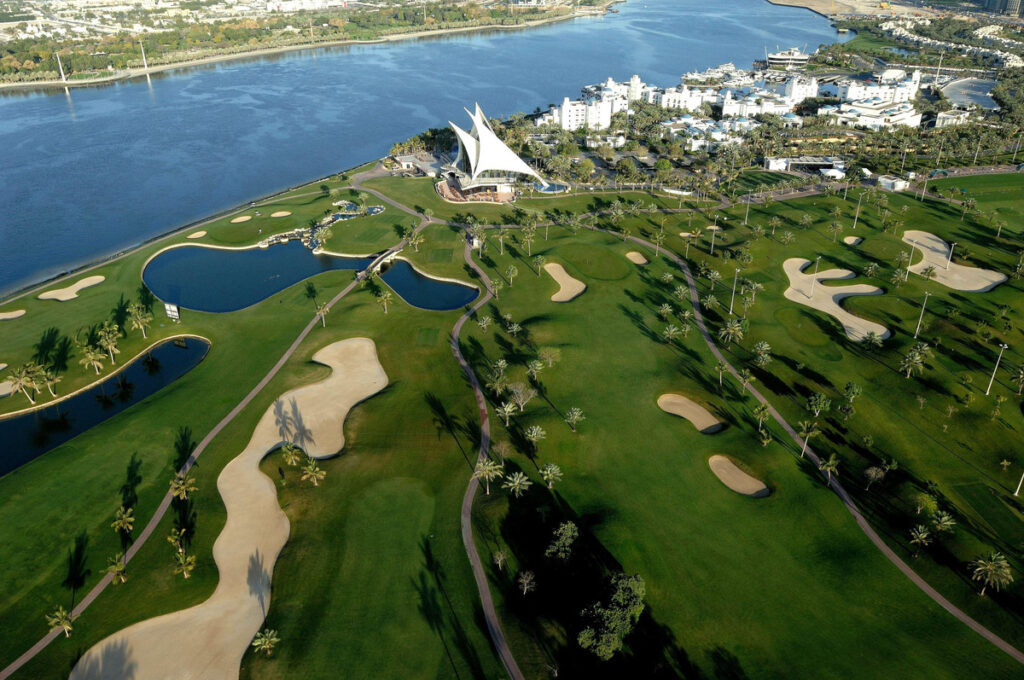 An aerial view of Dubai Creek Golf & Yacht Club, with green fairways, sand bunkers, the sail-shaped clubhouse and Park Hyatt Dubai beside the water.