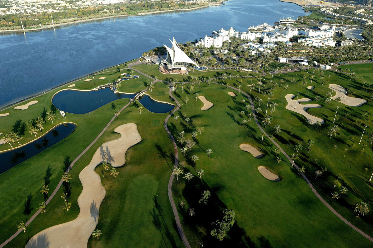 An aerial view of Dubai Creek Golf & Yacht Club, with green fairways, sand bunkers, the sail-shaped clubhouse and Park Hyatt Dubai beside the water.