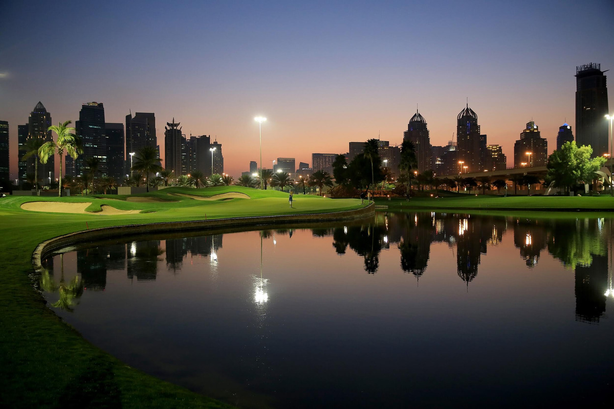 An illuminated green and sand bunkers beside a lake on the Faldo Course, with towers silhouetted against dusk.