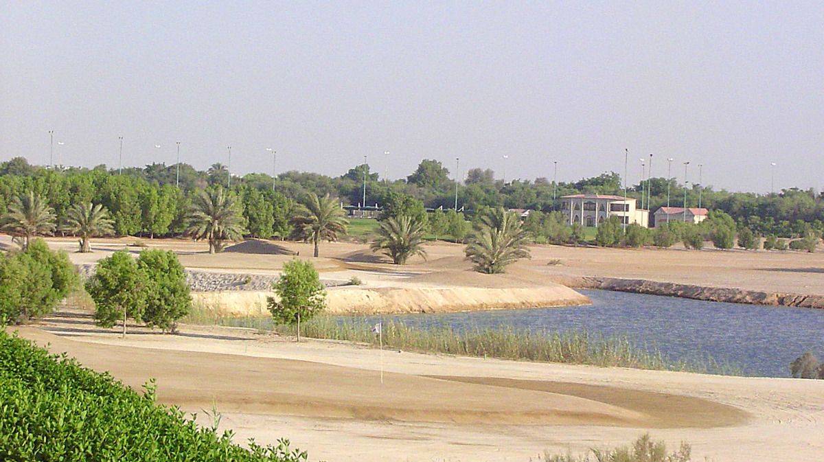 A flag stands on a brown sand putting surface beside water and palms at Al Ghazal Golf Club.