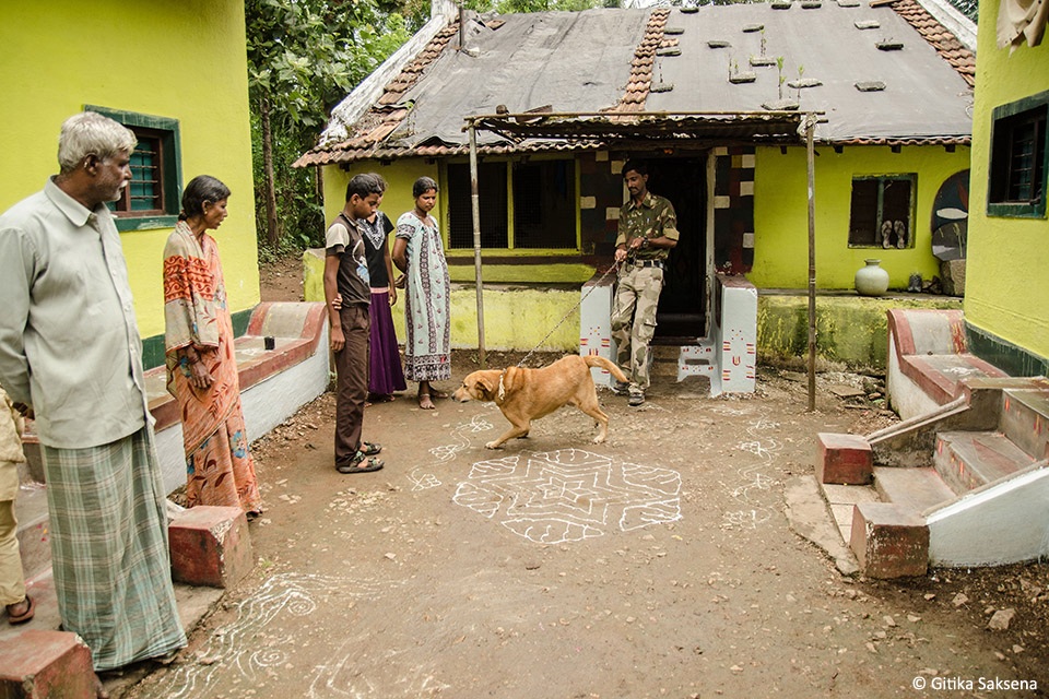 Somanna and his family stand in a courtyard with their dog; a younger man in naturalist uniform holds its lead.