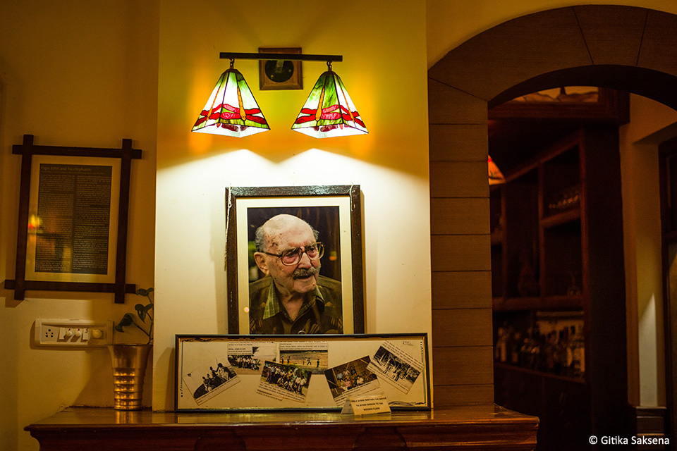 A framed photograph of John Wakefield and a display of clippings beneath two lamps inside Kabini River Lodge.