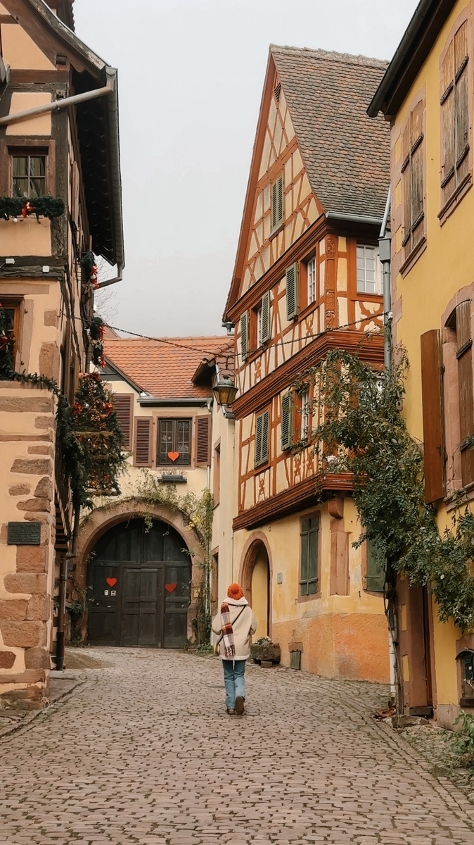 Olga in an orange hat and winter coat beside a Renaissance house and stone archway in Kaysersberg.