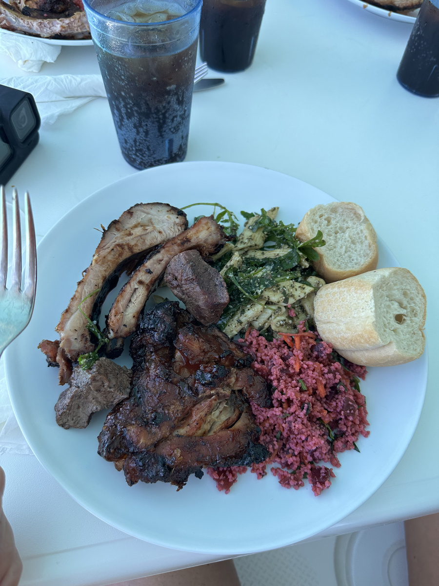 A plate of grilled meat with side dishes and bread, beside a glass with ice on the catamaran’s table.