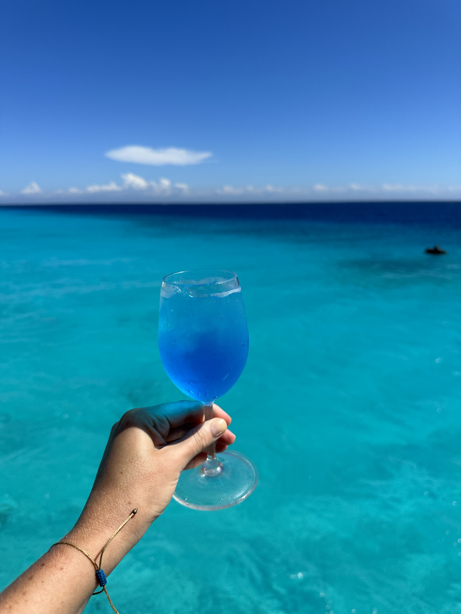 A hand holding a glass of blue liquid above turquoise water beside Klein Curaçao.