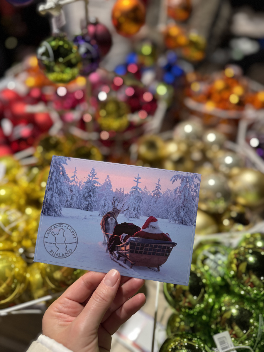 A hand holding a postcard of Santa in a reindeer sleigh, with colourful Christmas baubles in the shop behind it.