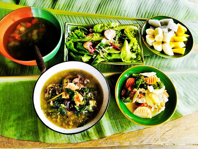 Soup, salad, cooked vegetables and sliced fruit arranged on banana leaves.