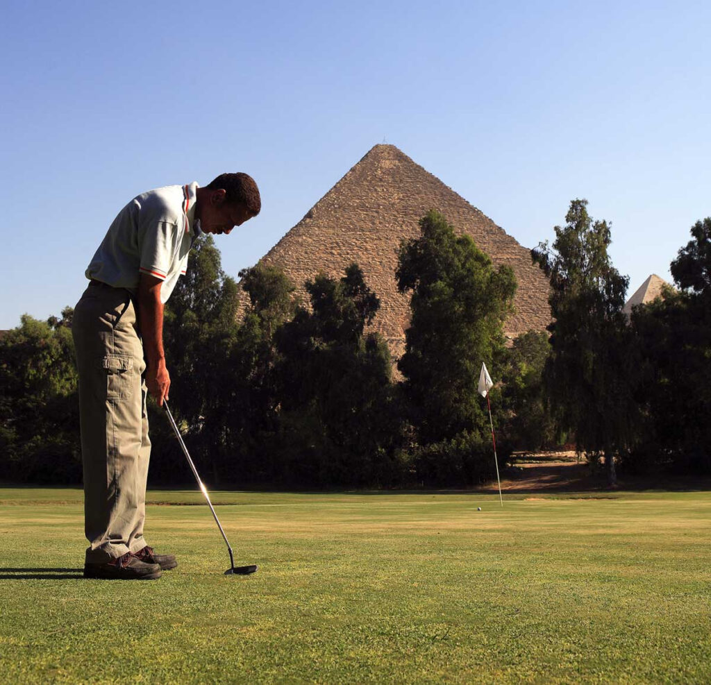 A golfer putts on a Mena House green with the Great Pyramid rising behind the trees.