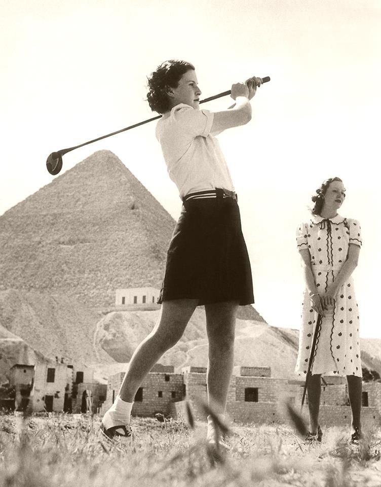 Two women with golf clubs in front of the Great Pyramid, one finishing a swing.
