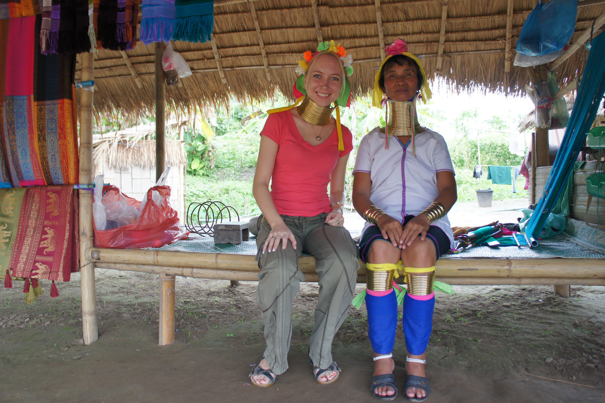 Olga wearing a brass collar and flowered headdress beside a Kayan woman at a village stall.