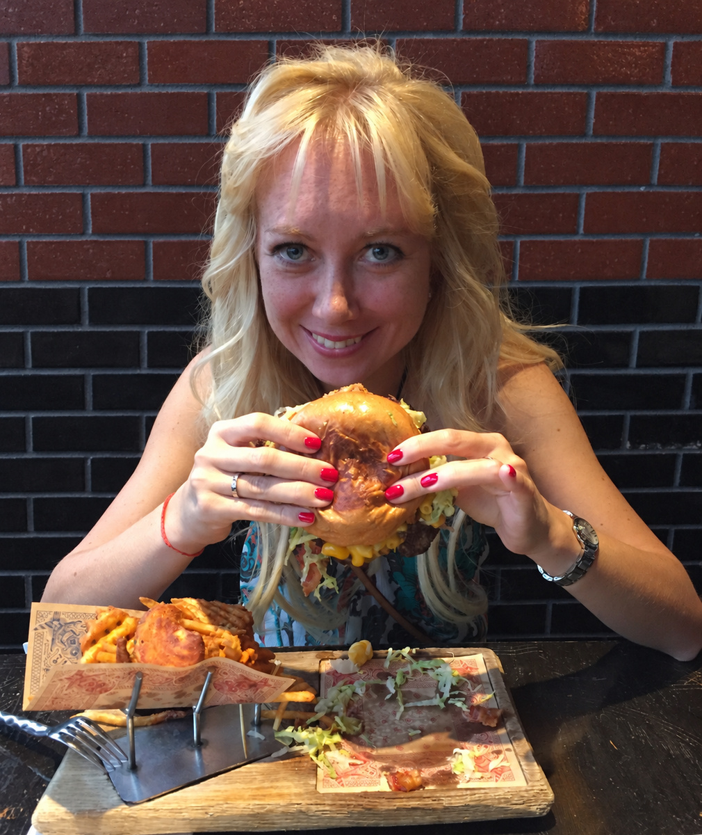 Olga smiling at the camera over a large burger with macaroni cheese, held in both hands against a brick wall.