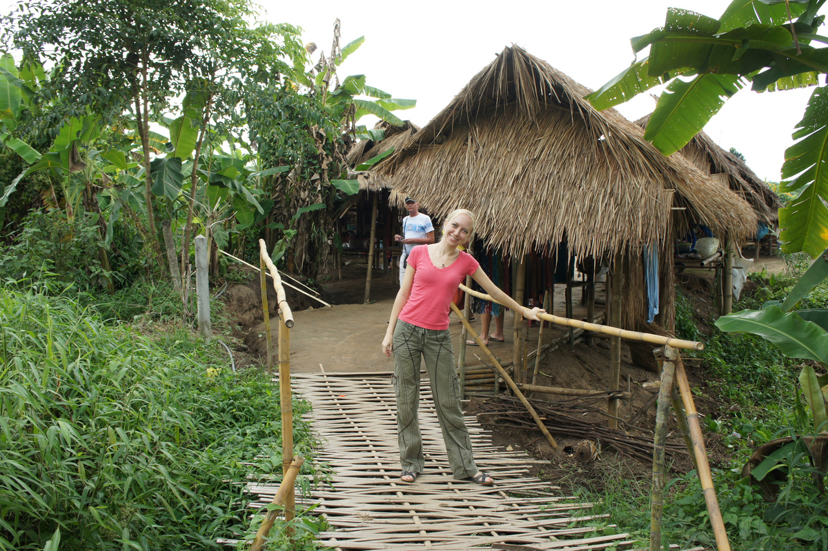 Olga standing on a narrow bamboo footbridge beside a thatched village hut.