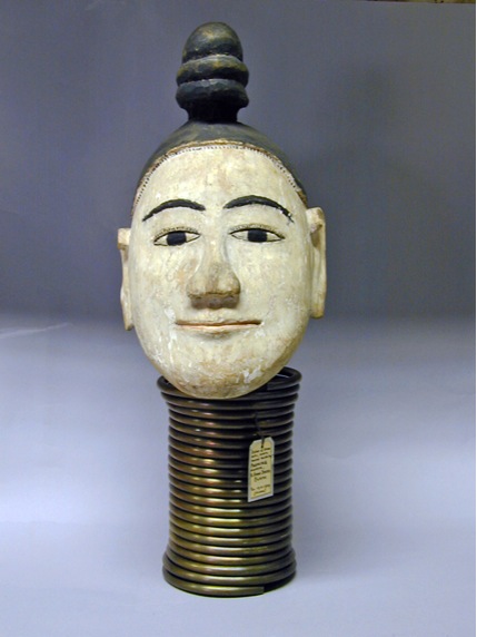 A carved and painted wooden woman’s head mounted above a tall brass neck coil.