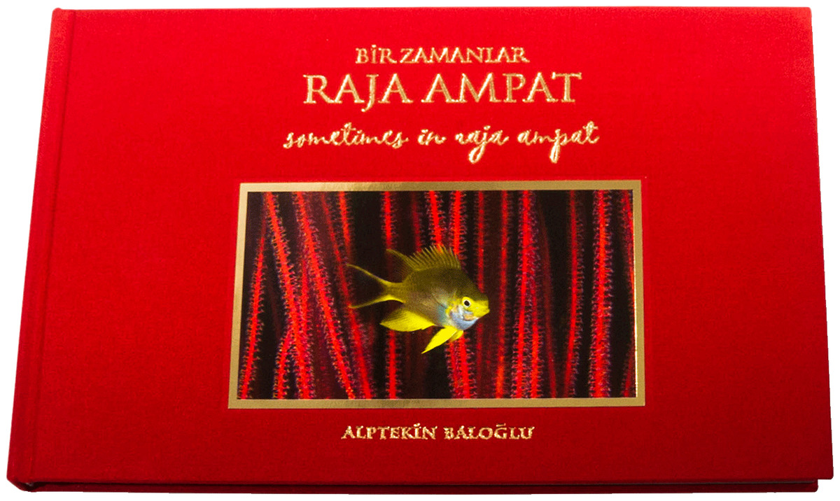 Alptekin Baloğlu’s Sometimes in Raja Ampat, a red landscape hardcover lettered in gold, with a yellow damselfish among red sea whips framed on the cover.