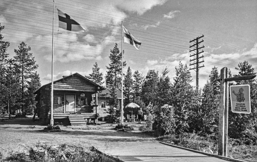 Black-and-white photograph of Roosevelt Cottage among trees, with two Finnish flags and an Arctic Circle sign beside a wooden footbridge.