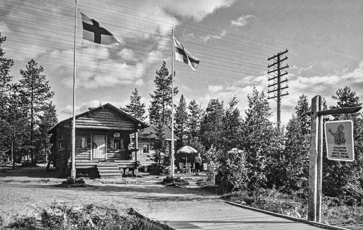 Black-and-white photograph of Roosevelt Cottage among trees, with two Finnish flags and an Arctic Circle sign beside a wooden footbridge.