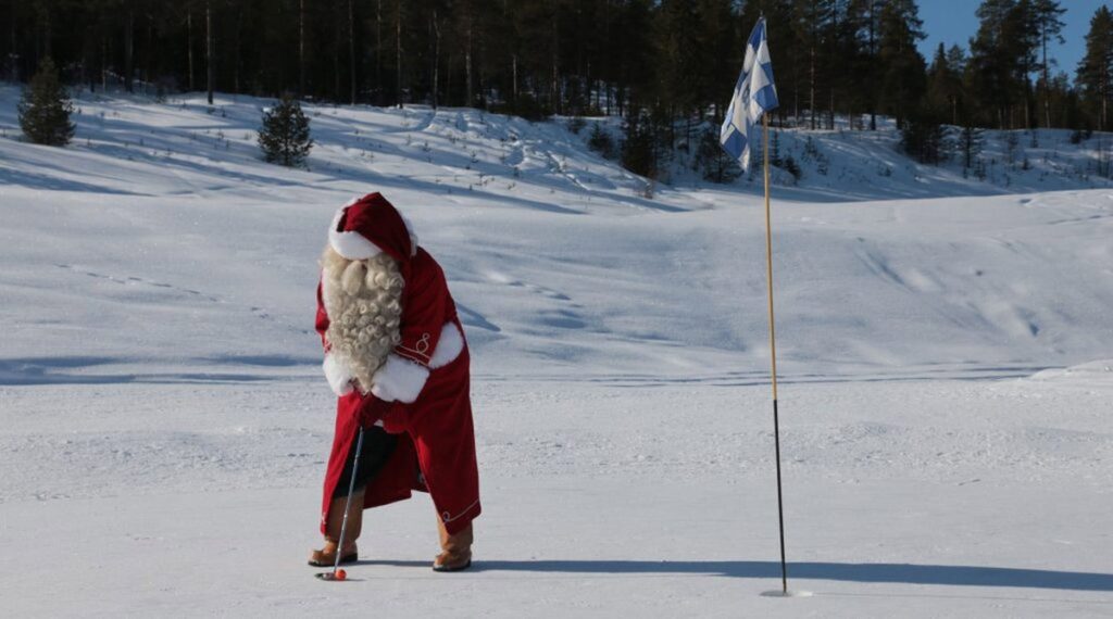 Santa Claus putting an orange golf ball on snow beside a Finnish flag.