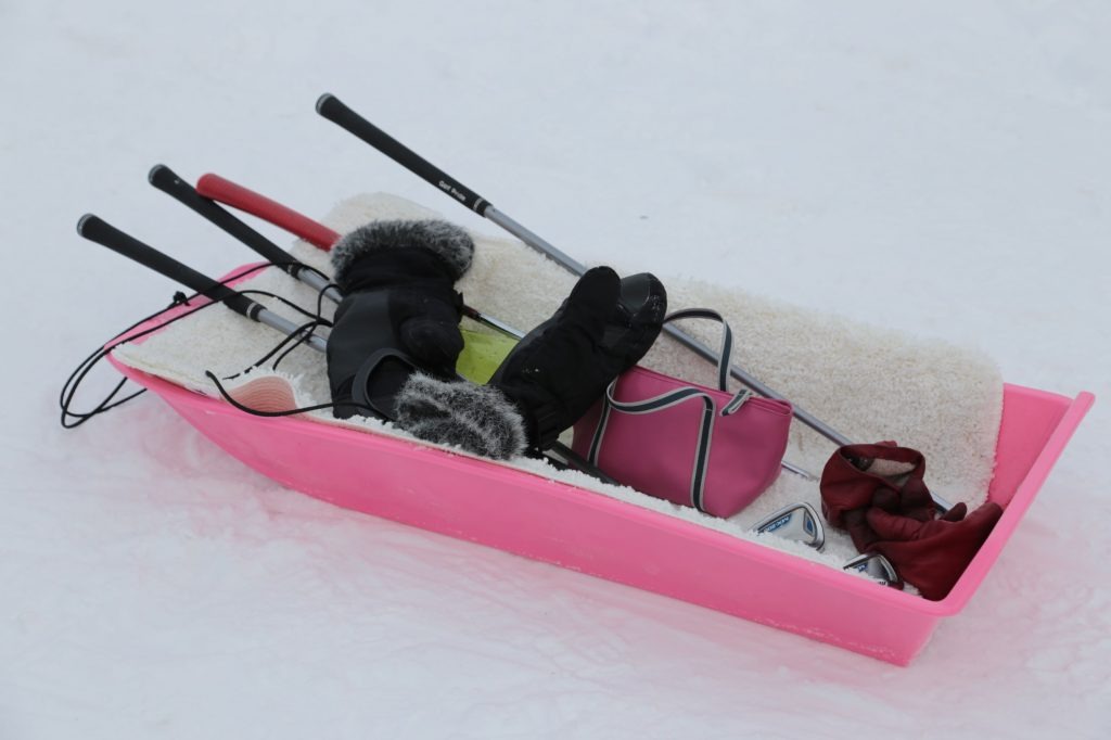 Golf clubs, mittens and a bag arranged in a pink sledge on snow.