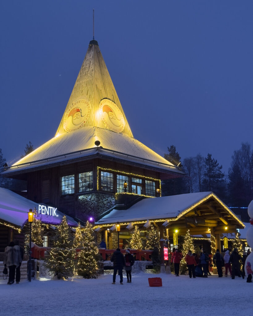 Illuminated Santa Claus Office tower above visitors on the snowy village square at dusk.