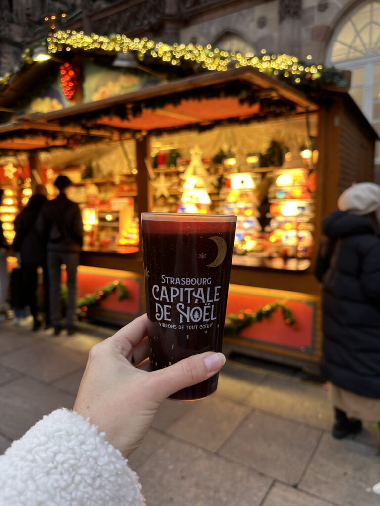 A cup bearing Strasbourg’s Capital of Christmas lettering held in front of an illuminated market stall.