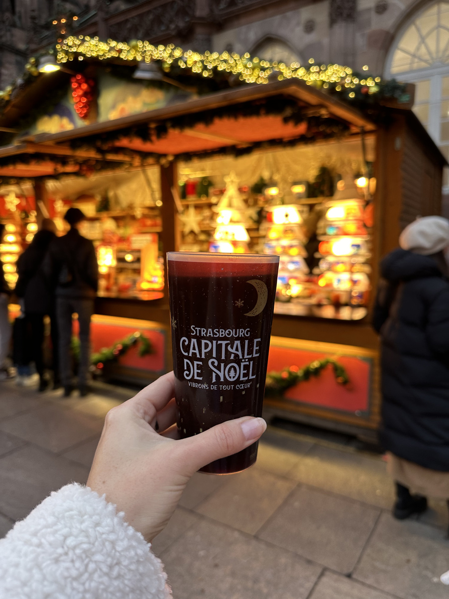 A cup bearing Strasbourg’s Capital of Christmas lettering held in front of an illuminated market stall.