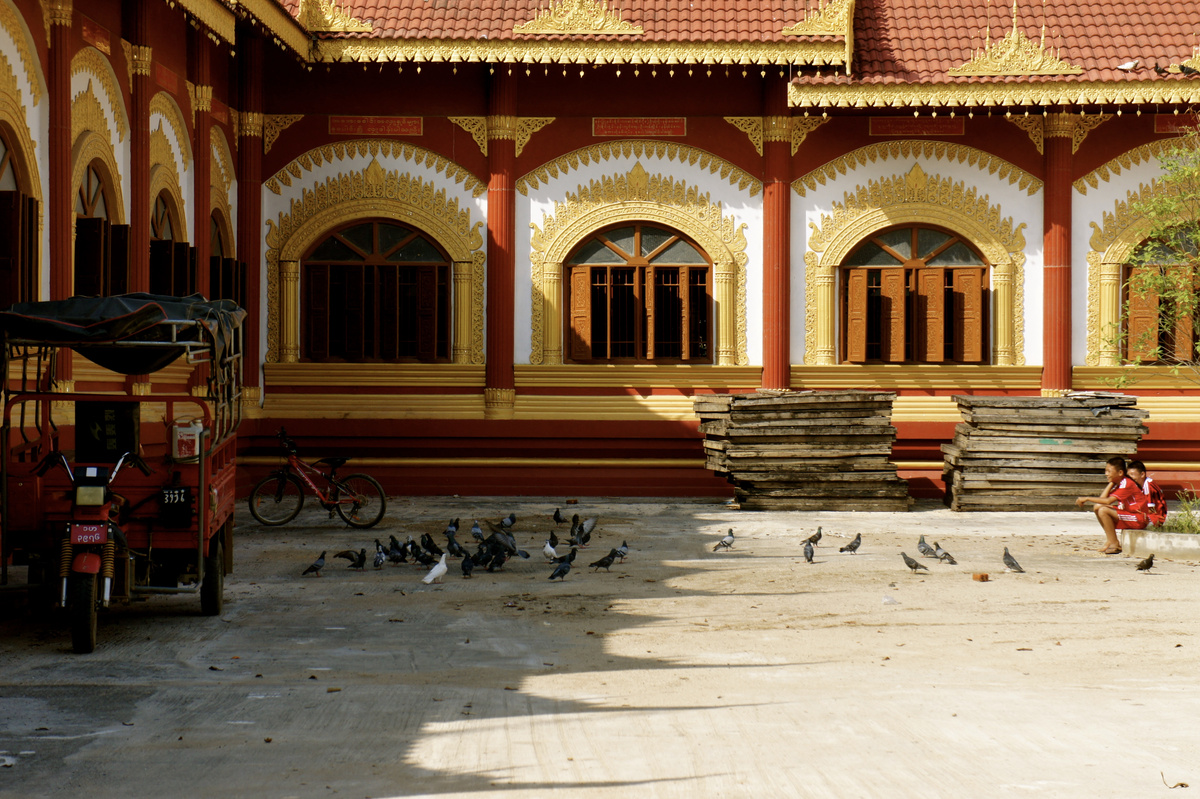 Pigeons and two children occupy a temple courtyard beside red columns and gilded window arches.
